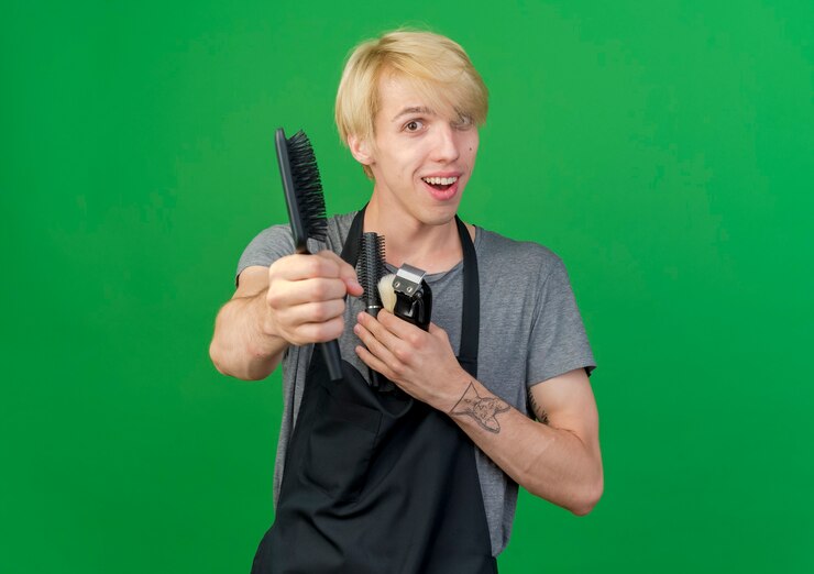 Foto grátis barbeiro profissional com avental segurando o aparador e escovas de cabelo oferecendo uma escova para a câmera sorrindo
