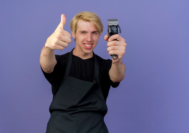 Barbeiro profissional com avental segurando o aparador, feliz e alegre, mostrando os polegares em pé sobre a parede azul