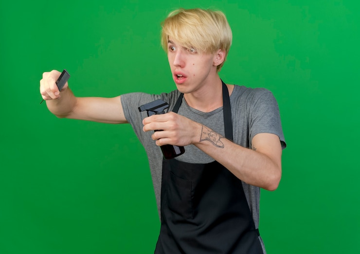 Foto grátis barbeiro profissional com avental segurando um pente de cabelo e spray, parecendo de lado surpreso e confuso