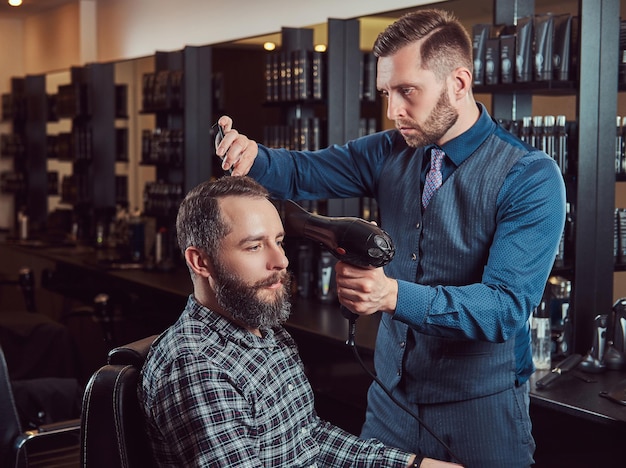 Barbeiro profissional trabalhando com um cliente em um salão de cabeleireiro, usa um secador de cabelo. corte de cabelo masculino em uma barbearia.