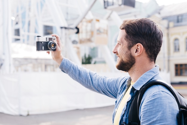 Barbudo homem sorridente tomando selfie com câmera retro durante a viagem