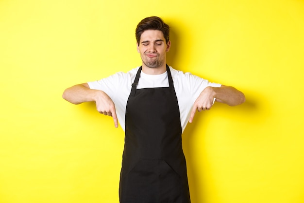 Barista duvidoso reclamando, apontando o dedo para baixo e fazendo uma careta de descontentamento, em pé com avental preto sobre fundo amarelo.