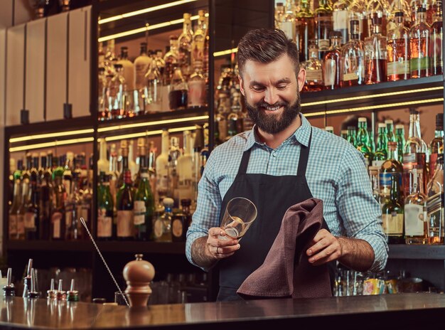 Barman brutal elegante e alegre está limpando um copo com um pano no fundo do balcão de bar.