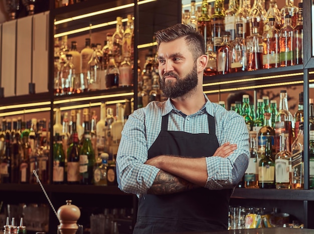 Barman brutal elegante em uma camisa e avental em pé com os braços cruzados no fundo do balcão de bar.