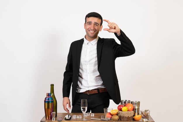 Barman masculino com vista frontal em um terno clássico em pé em frente à mesa com bebidas na parede branca bar de bebidas alcoólicas no bar noturno masculino