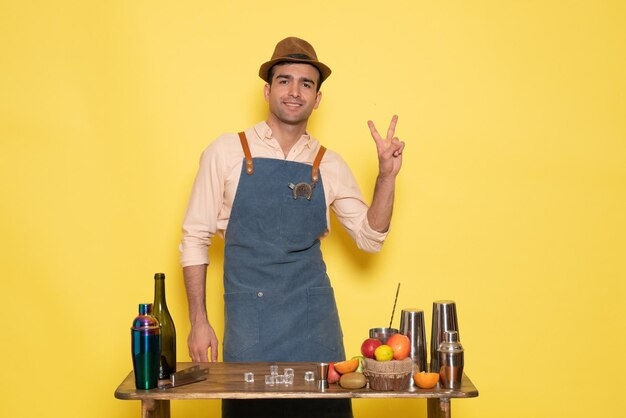 Barman masculino de frente para a mesa com bebidas e frutas sorrindo na parede amarela.