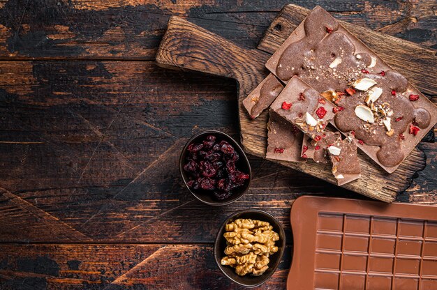 Barra de chocolate ao leite artesanal caseiro com avelãs, amendoim, cranberries e framboesas liofilizadas. fundo de madeira escuro. vista do topo. copie o espaço.