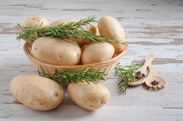 Batatas cruas em uma cesta de vime com folhas de alecrim natural em uma mesa de madeira rústica