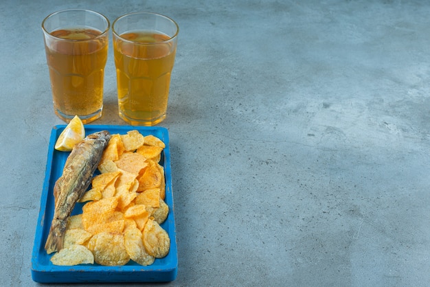 Batatas fritas e peixe num prato de madeira ao lado de dois copos de cerveja, na mesa de mármore.