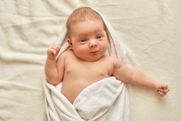 Bebé depois do banho, deitado na cama no cobertor branco, olhando para a câmera, sendo enrolado em uma toalha, bebê depois do banho, encantador garoto recém-nascido.