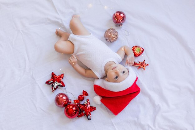 Bebê em uma roupa branca e um chapéu de papai noel está deitado de costas em um lençol branco cercado por brinquedos vermelhos de árvore de natal. inverno, ano novo. espaço para texto. foto de alta qualidade