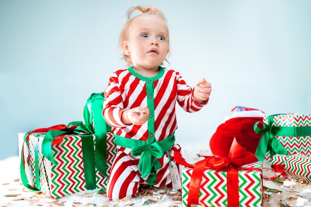 Bebê fofo menina de 1 ano de idade perto de chapéu de Papai Noel posando sobre fundo de Natal. Sentado no chão com uma bola de Natal. Temporada de férias.