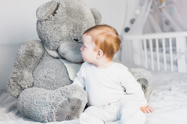 Bebê loiro fofo na cama branca com ursinho de pelúcia