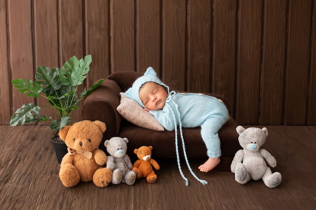 Bebê recém-nascido pouco agradável e bonito menino dormindo no pequeno sofá marrom de pijama azul, rodeado por ursos de plantas e brinquedos