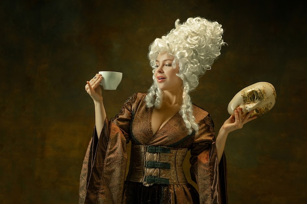 Beber café com máscara. Retrato de uma jovem medieval em roupas vintage marrons na parede escura. Modelo feminino como duquesa, pessoa real. Conceito de comparação de eras, moderno, moda.