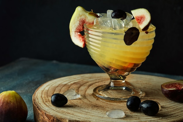 Bebida amarelo-laranja com figos e uvas em um carrinho de madeira no escuro