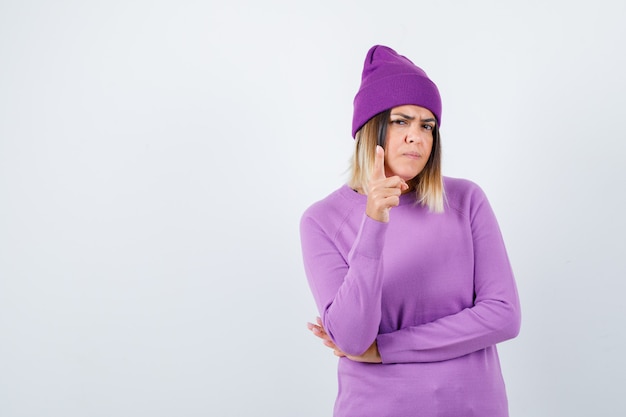 Bela dama apontando para cima em suéter, gorro e olhando confiante, vista frontal.