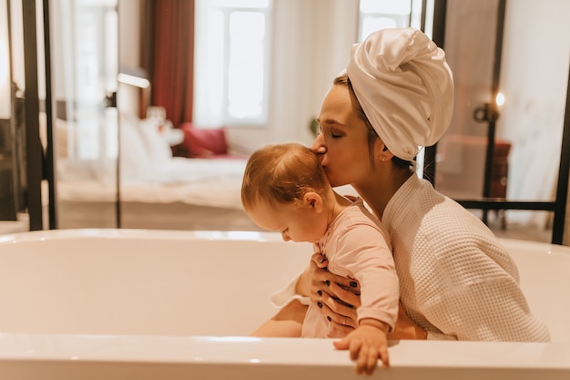 Foto grátis bela dama com roupa branca em casa beija seu filho. mãe e bebê estão sentados no banheiro.