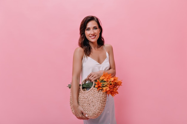 Bela dama de blusa branca contém um saco de palha e flores de laranja.  mulher bonita com roupa elegante leve segura bolsa de malha com buquê.
