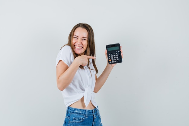 Bela dama de blusa branca, jeans, apontando para a calculadora e olhando alegre, vista frontal.