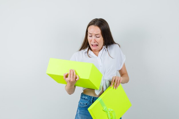 Bela dama de blusa branca, jeans, olhando para dentro da caixa amarela e parecendo enojada, vista frontal.