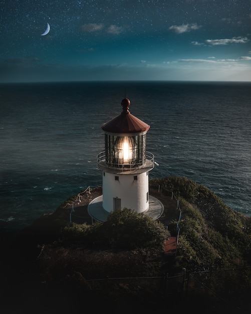 Bela foto aérea de um farol aceso em uma colina verde com a meia-lua no céu noturno