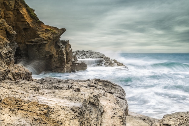 Bela foto das ondas do mar batendo nas rochas à beira-mar com um céu nublado e cinza