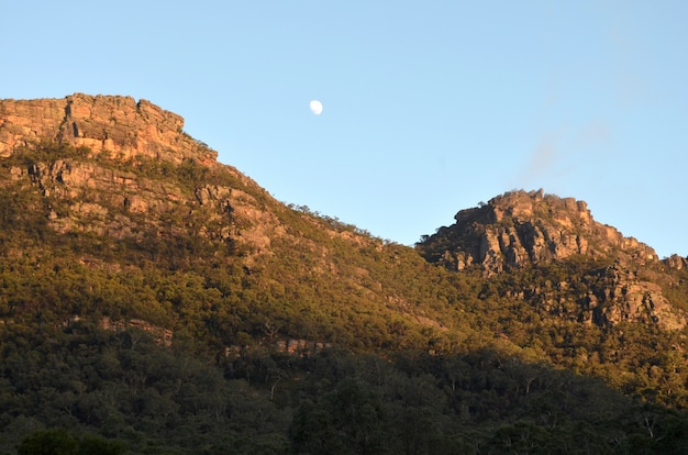 Foto grátis bela foto de montanhas arborizadas sob um céu claro, com uma lua visível durante o dia