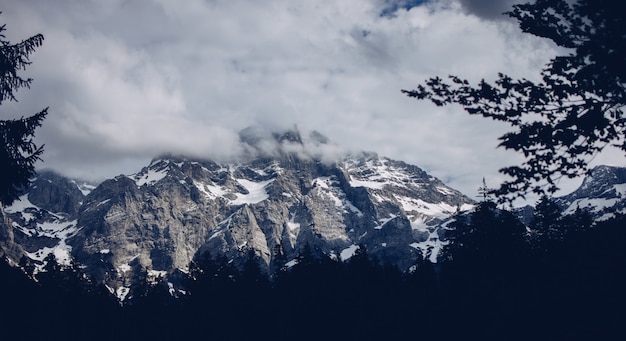 Bela foto de montanhas rochosas e nevadas com incríveis nuvens e vegetação ao redor