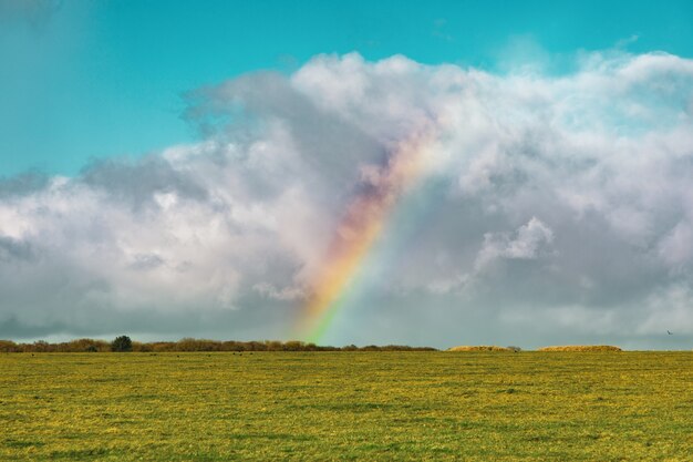 Bela foto de um campo gramado vazio com um arco-íris à distância sob um céu azul nublado