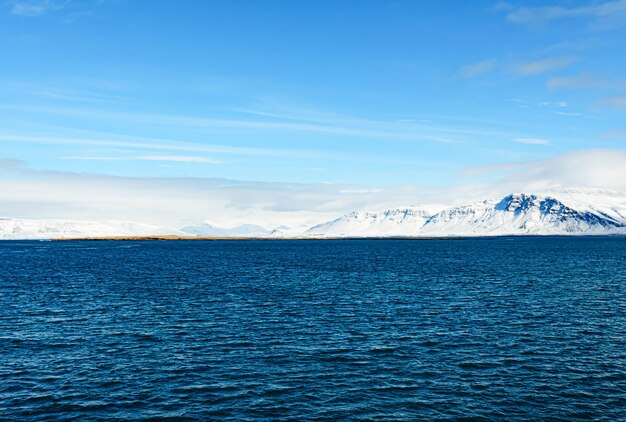 Bela foto de um mar azul cheio de ondas em frente a uma montanha de neve na Islândia