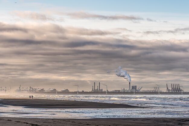 Bela foto de um mar com moinhos de vento e uma fábrica ao longe, sob um céu nublado