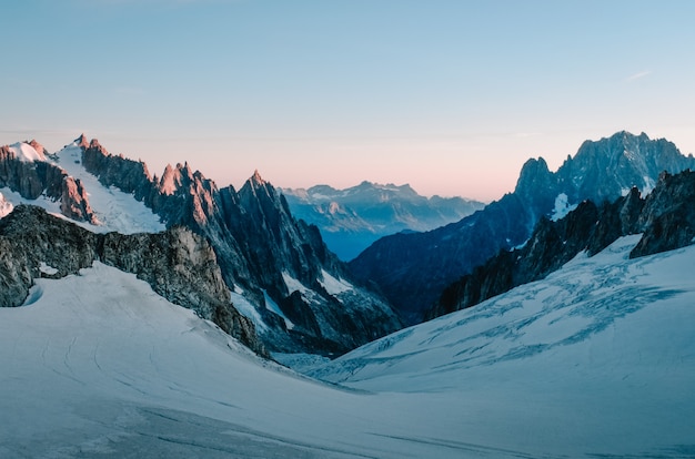 Bela foto de uma colina de neve rodeada por montanhas com o céu rosa claro