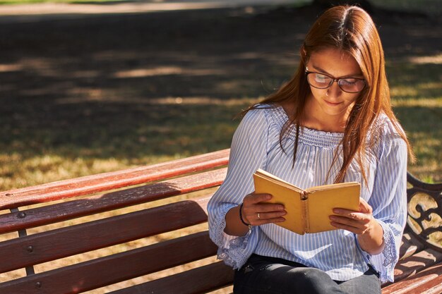 Bela foto de uma garota de camisa azul e óculos lendo um livro no banco