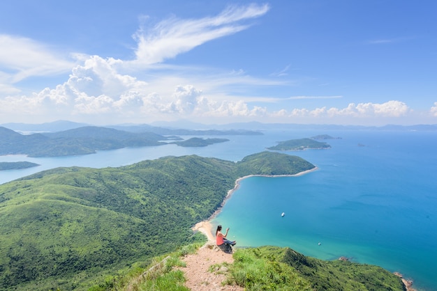 Bela foto de uma mulher sentada em um penhasco com uma paisagem de colinas arborizadas e um oceano azul