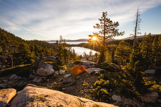 Bela foto de uma tenda laranja na montanha rochosa, rodeada por árvores durante o pôr do sol