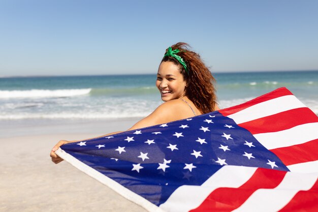 Bela jovem acenando a bandeira americana na praia ao sol