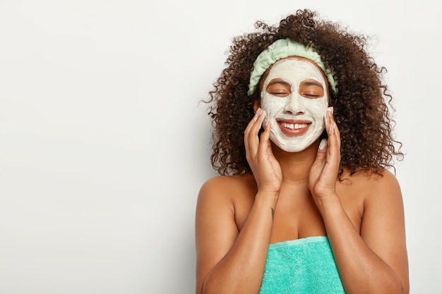 Foto grátis bela jovem aplica máscara facial de argila branca, faz tratamentos de rejuvenescimento em salão de spa, usa bandana, fica com toalha macia turquesa em volta do corpo nu, limpa a pele