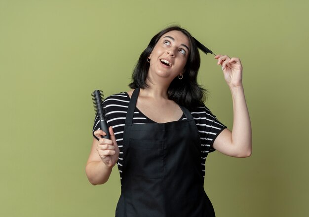 Foto grátis bela jovem cabeleireira com avental segurando escovas de cabelo e sorrindo alegremente com o rosto feliz em pé sobre a parede de luz