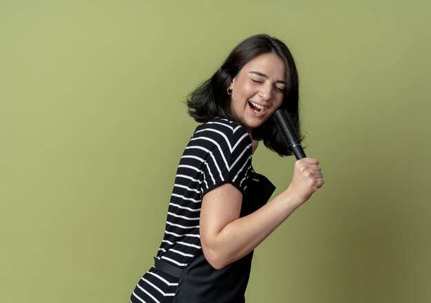 Bela jovem cabeleireira de avental segurando escovas de cabelo, sorrindo alegremente com uma cara feliz, usando escova de cabelo como microfone cantando em pé sobre a parede de luz