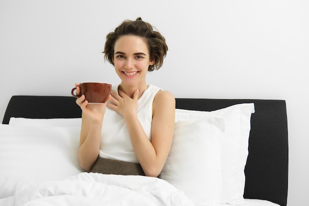 Foto grátis bela jovem com uma xícara de café bebendo cappuccino na cama e sorrindo