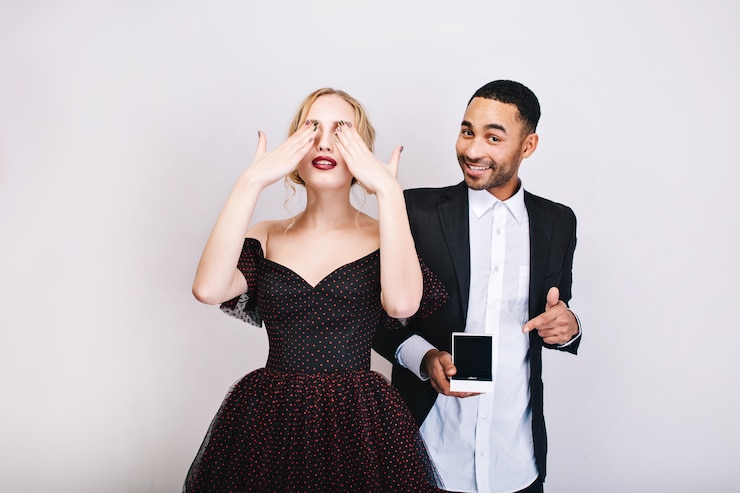 Foto grátis bela jovem com vestido de noite de luxo fecha os olhos com as mãos, esperando a surpresa do cara bonito com anel, sorrindo por trás. dia dos namorados, proposta, amantes.