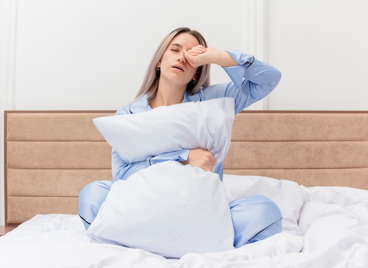 Foto grátis bela jovem de pijama azul sentada na cama com travesseiro, acordando com cansaço matinal no interior do quarto