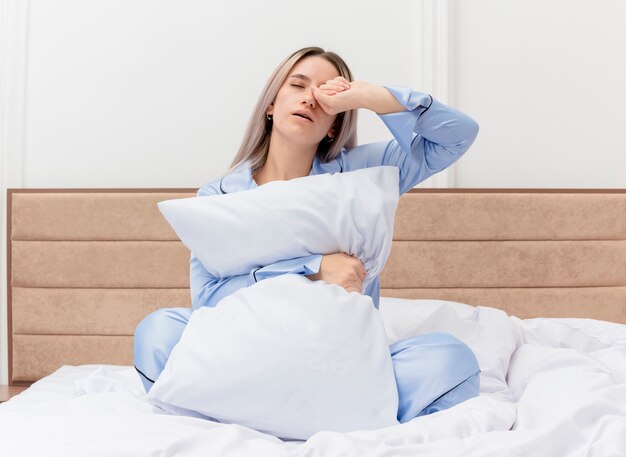 Bela jovem de pijama azul sentada na cama com travesseiro, acordando com cansaço matinal no interior do quarto