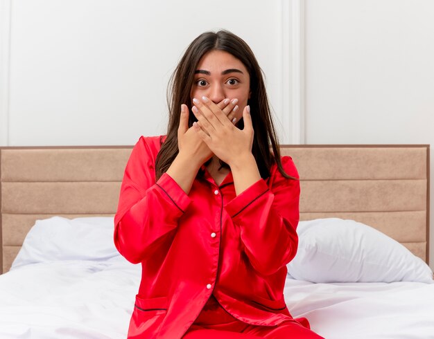 Bela jovem de pijama vermelho sentada na cama, olhando para a câmera, maravilhada e surpresa, cobrindo a boca com as mãos no interior do quarto sobre fundo claro