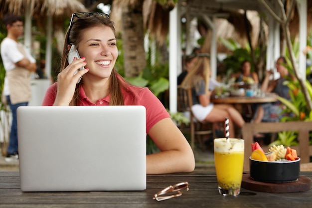 Bela jovem empresária falando no celular com um olhar feliz, sentado à mesa de madeira com um coquetel e aberto o computador laptop genérico