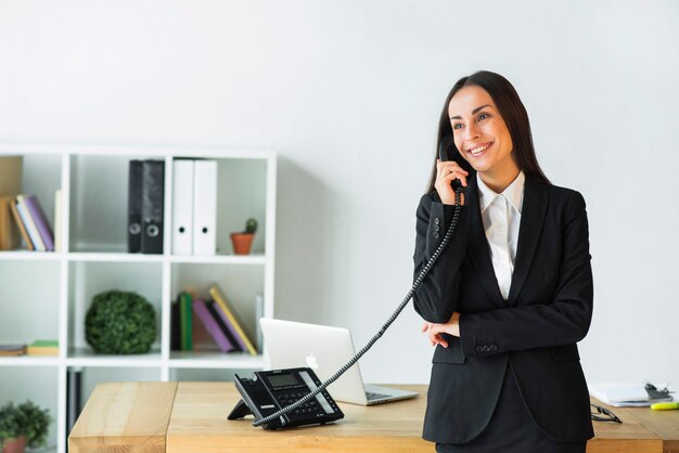 Bela jovem empresária falando no telefone em pé perto da mesa de madeira