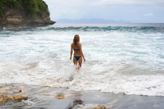 Bela jovem esbelta com longos cabelos loiros em um maiô na praia perto do oceano. Relaxe na praia. Férias tropicais. Uma mulher entra na água para nadar.