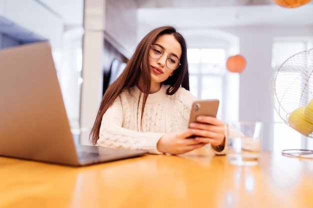 Bela jovem está sentado na cozinha com laptop com videocall no telefone