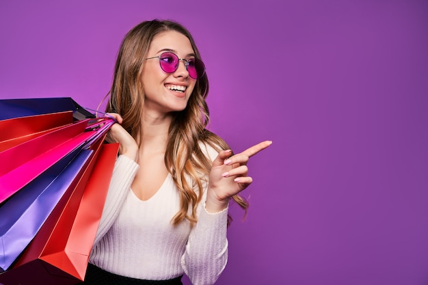 Bela jovem loira sorridente apontando em óculos de sol segurando sacolas de compras e um cartão de crédito em uma parede rosa